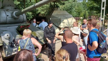 Grupa osób słucha opowieści mężczyzny ubranego w mundur czołgisty. W tle czołg i ciągnik opancerzony