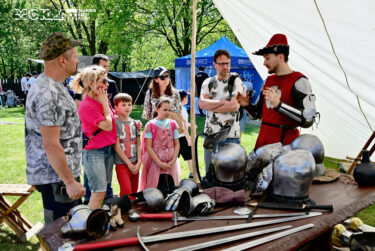 Na pierwszym planie stół z elementami zbroi rycerskiej - mieczami i chełmami. W tle mężczyzna ubrany w srój średniowieczny mówi coś do stojącej przed nim grupy osób.