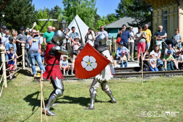 Dwóch walczących rycerzy w strojach ze średniowieca. Walczą na miecze. Jest piękna słoneczna pogoda. W tle ludzie przyglądający się walce.