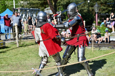 Dwóch walczących rycerzy w strojach średniowiecznych. Walczą na miecze. W tle grupa obserwujących osób.