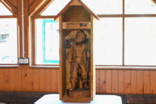 Drewniana kapliczka/rzeźba przedstawiająca wizerunek rybaka z wędkarskimi atrybutami.